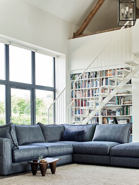 Blue Alexander and James Summerton corner sofa with footstool in a white open plan living room with a large built in bookcase